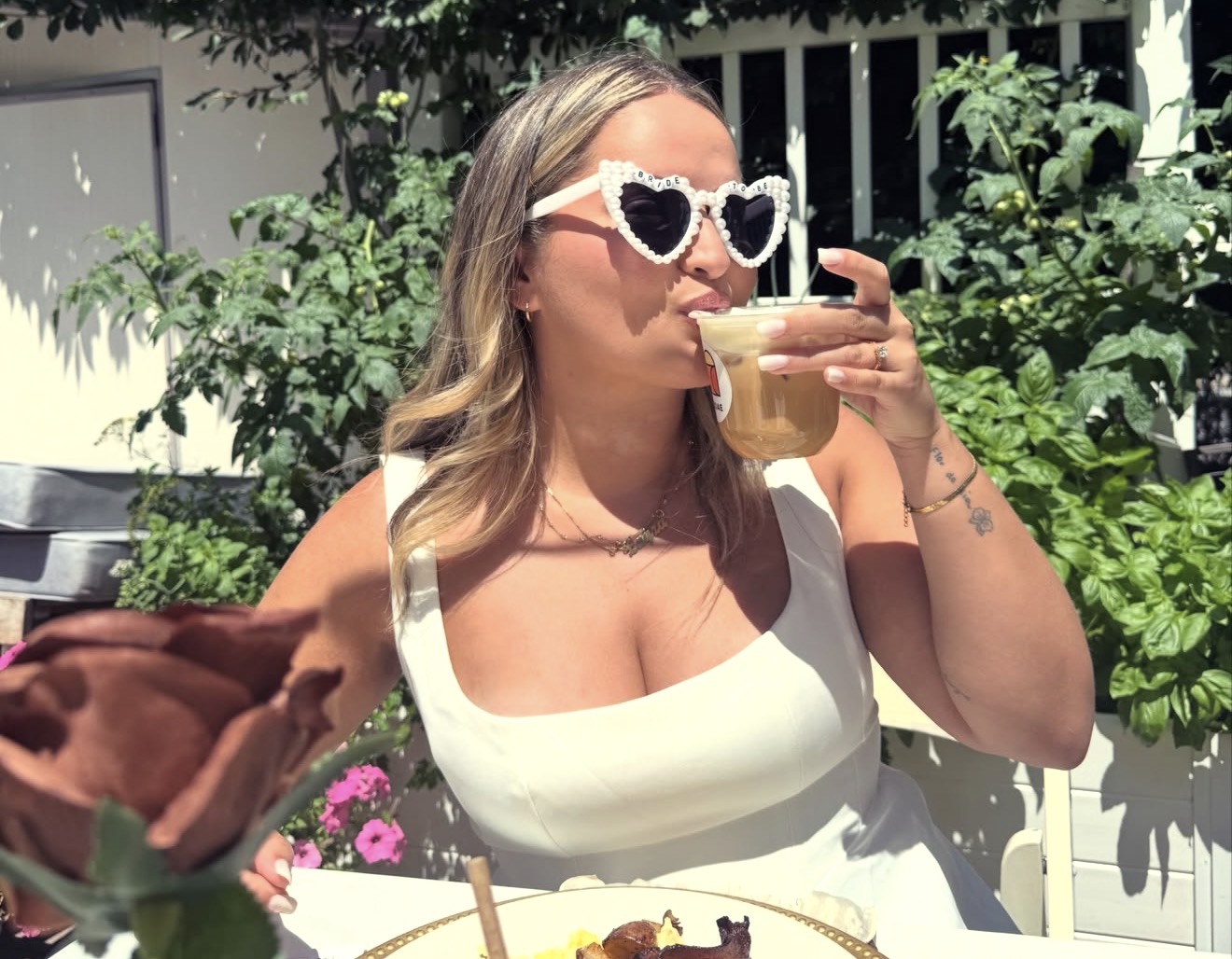 Layza in white with bride-to-be heart sunglasses sipping a drink in the garden