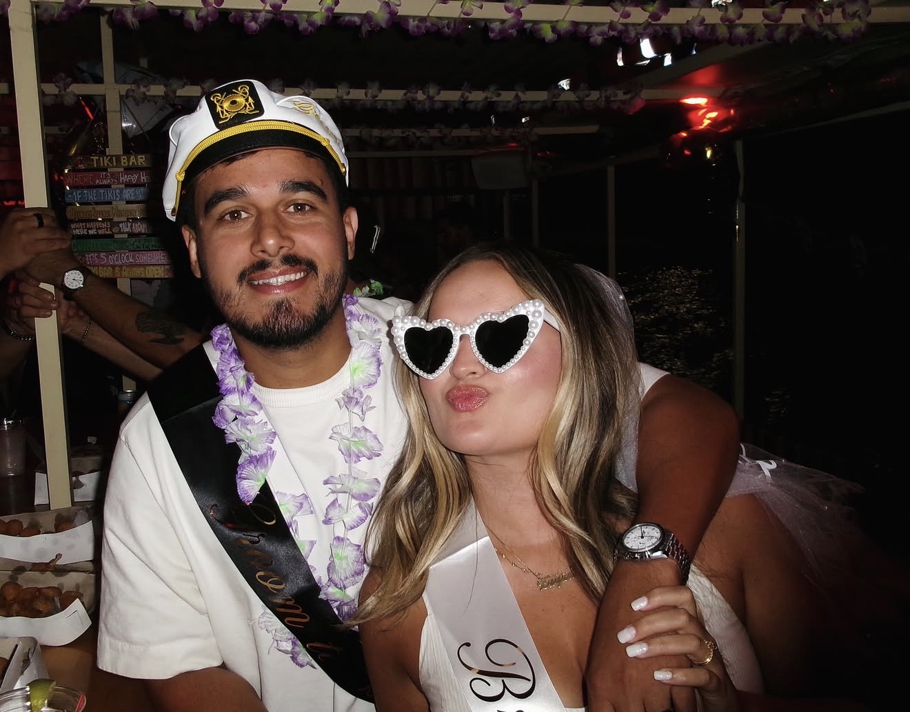 The bride and groom in sashes, captain's hat and heart glasses at the tiki bar