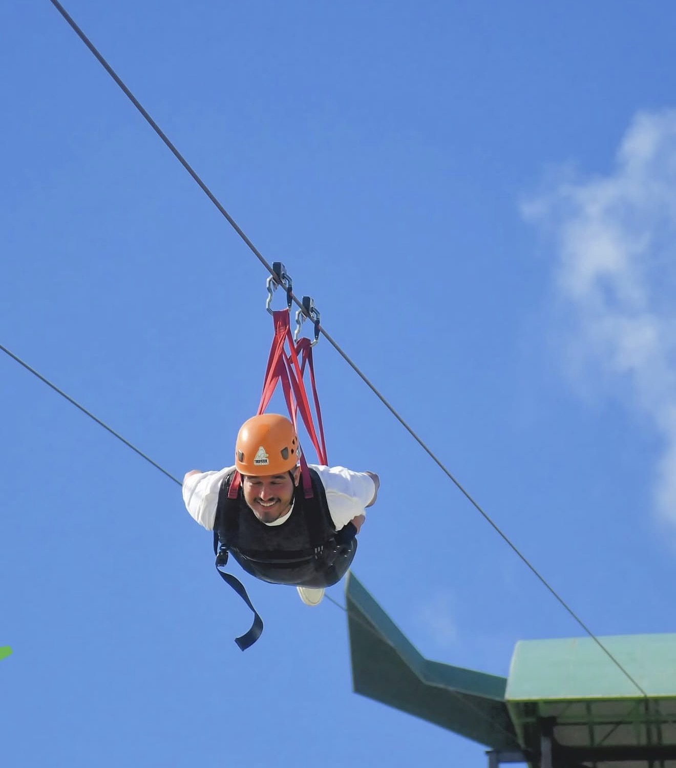 Matheus grinning mid-flight on a zipline against blue sky